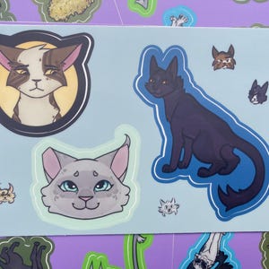May include: A sheet of cat-themed stickers with various cat illustrations. The stickers include a brown and white cat in a circular frame, a black cat, and a grey cat with blue eyes. Additional small cat graphics are also present.