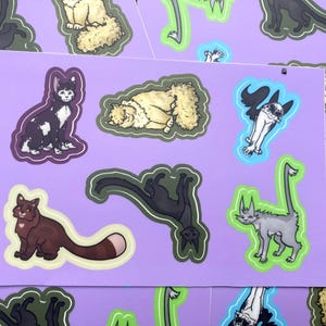 May include: A sheet of cat stickers featuring diverse cat illustrations. The stickers depict cats in various poses and colours, including black, brown, grey, and cream. Each sticker has a white outline and is set against a lilac background.