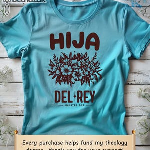 May include: A turquoise blue t-shirt with the text "HIJA DEL REY - GALATAS 3:26" in brown lettering. The text is surrounded by a floral design.