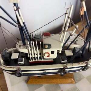 May include: A detailed wooden model of a fishing boat. The boat has a white hull with blue stripes, a white cabin, and black accents. The model includes rigging, masts, and a wooden base. A small life preserver is visible.