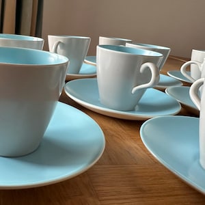 Puede incluir: Un juego de tazas de té de cerámica blanca con interior azul claro, sobre platillos a juego. Las tazas tienen un asa curvada y un diseño sencillo y elegante. Los platillos son ovalados.