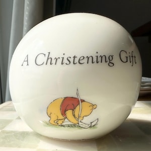 May include: A white, spherical decorative item with the text "A Christening Gift" in black. A cartoon illustration of Winnie the Pooh is featured below the text, wearing a red shirt and yellow body.