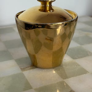 May include: A gold-colored, lidded container with a bulbous body and a small, round handle on top. The container has a geometric pattern and a shiny finish. The lid fits snugly on the container. The container is likely for storing sugar or other small items.