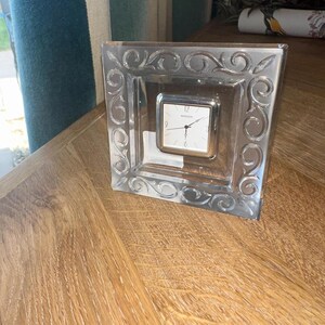 May include: A square, clear glass clock with a silver-toned clock face. The clock is set within a decorative, etched glass frame with swirling patterns. The clock sits on a wooden surface.