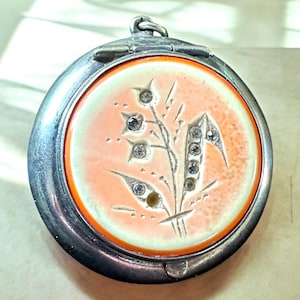 May include: A silver locket with a hinged lid. The locket has a peach-coloured enamel background with a white floral design and rhinestones.