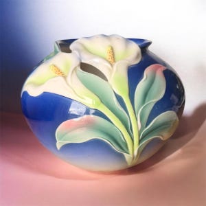 May include: A rounded ceramic vase featuring a deep blue gradient background with raised, three-dimensional white calla lily flowers. The design includes soft green leaves with subtle pink highlights, creating a textured floral relief on the surface.
