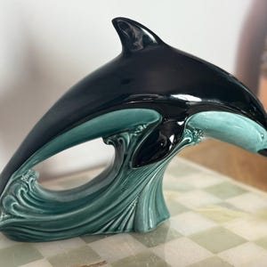 Könnte beinhalten: Keramik-Delfinfigur in glänzendem Schwarz und Türkis. Der Delfin springt über eine türkisfarbene Wellenbasis. Die Figur wird auf einer karierten Oberfläche präsentiert.