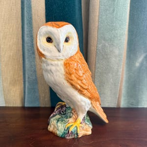 Può includere: Una statuetta di gufo in ceramica con viso bianco, corpo arancione e zampe gialle. Il gufo è appollaiato su una base di roccia colorata. La statuetta è ambientata su uno sfondo di tenda a strisce.