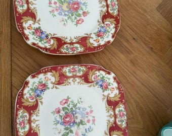 May include: Two square, white plates with a red border and floral design. The plates have a central bouquet of pink, blue, and yellow flowers. The red border is decorated with gold accents and floral patterns, creating a vintage aesthetic.