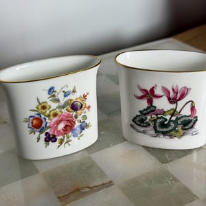 May include: Two white porcelain vases with gold rims. One vase features a floral design with pink roses, blue and yellow flowers. The other vase has a pink flower and green leaf design.