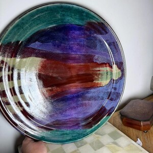 May include: A large, round ceramic plate with a hand-painted design. The plate features a teal green rim, a central area of purple and red hues, and a white and tan stripe pattern. The plate is held by a hand.