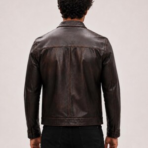 May include: A dark brown leather jacket with a distressed finish, featuring a classic collar, long sleeves with buttoned cuffs, and a straight hemline. The jacket is worn over black trousers, showcasing a structured back panel design.