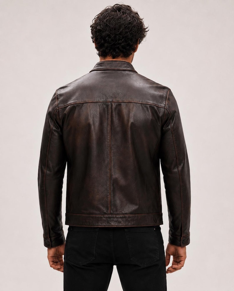 May include: A dark brown leather jacket with a distressed finish, featuring a classic collar, long sleeves with buttoned cuffs, and a straight hemline. The jacket is worn over black trousers, showcasing a structured back panel design.