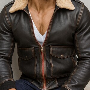 Puede incluir: Chaqueta bomber de cuero marr&oacute;n con cuello de borrego beige. La chaqueta tiene dos bolsillos delanteros, una cremallera roja y pu&ntilde;os acanalados. La chaqueta se lleva sobre una camisa blanca.