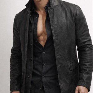 May include: Black leather jacket with a button-up front and two front pockets. The jacket is worn over a black button-down shirt. The collar of the jacket is a stand-up style.