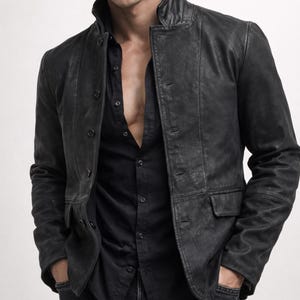May include: Black leather jacket with a button-up front, worn over a black button-down shirt. The jacket has a notched collar and two front pockets. The outfit is completed with black jeans.