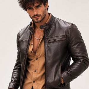 May include: A dark brown leather jacket with a stand-up collar, snap button closure, and a zippered chest pocket. The jacket is worn over a tan button-down shirt. A silver chain necklace is visible around the neck, and a silver bracelet is on the wrist.