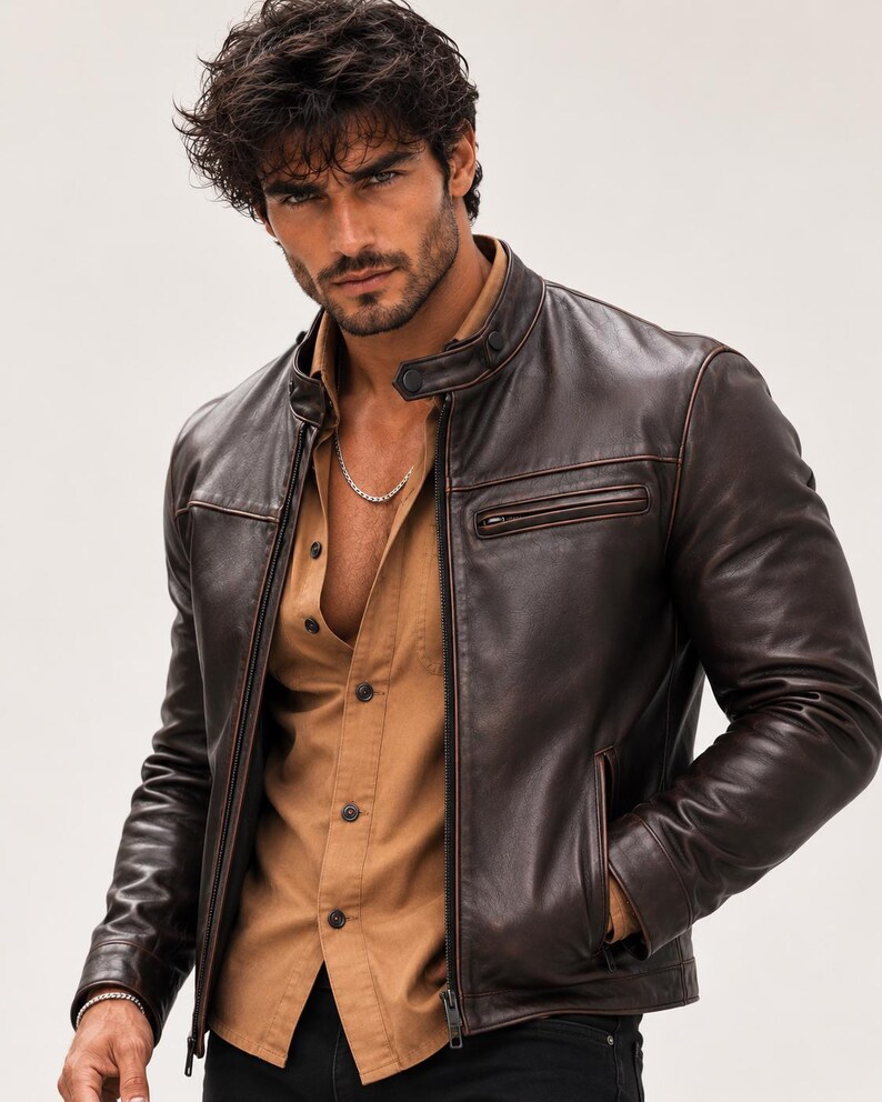 May include: A dark brown leather jacket with a stand-up collar, snap button closure, and a zippered chest pocket. The jacket is worn over a tan button-down shirt. A silver chain necklace is visible around the neck, and a silver bracelet is on the wrist.