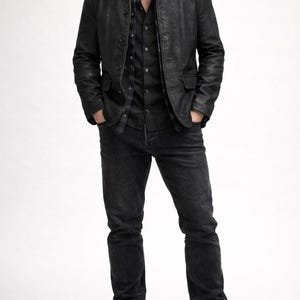 May include: A black leather jacket with a button-up front, paired with a black button-down shirt and dark wash jeans. The outfit is completed with black leather boots. The overall style is a classic, monochromatic look.