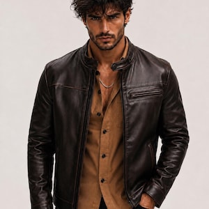 May include: A dark brown leather jacket with a stand-up collar, a front zip fastening, and a zipped chest pocket. The jacket is worn over a tan button-down shirt and black trousers, featuring a silver chain necklace around the neck area.