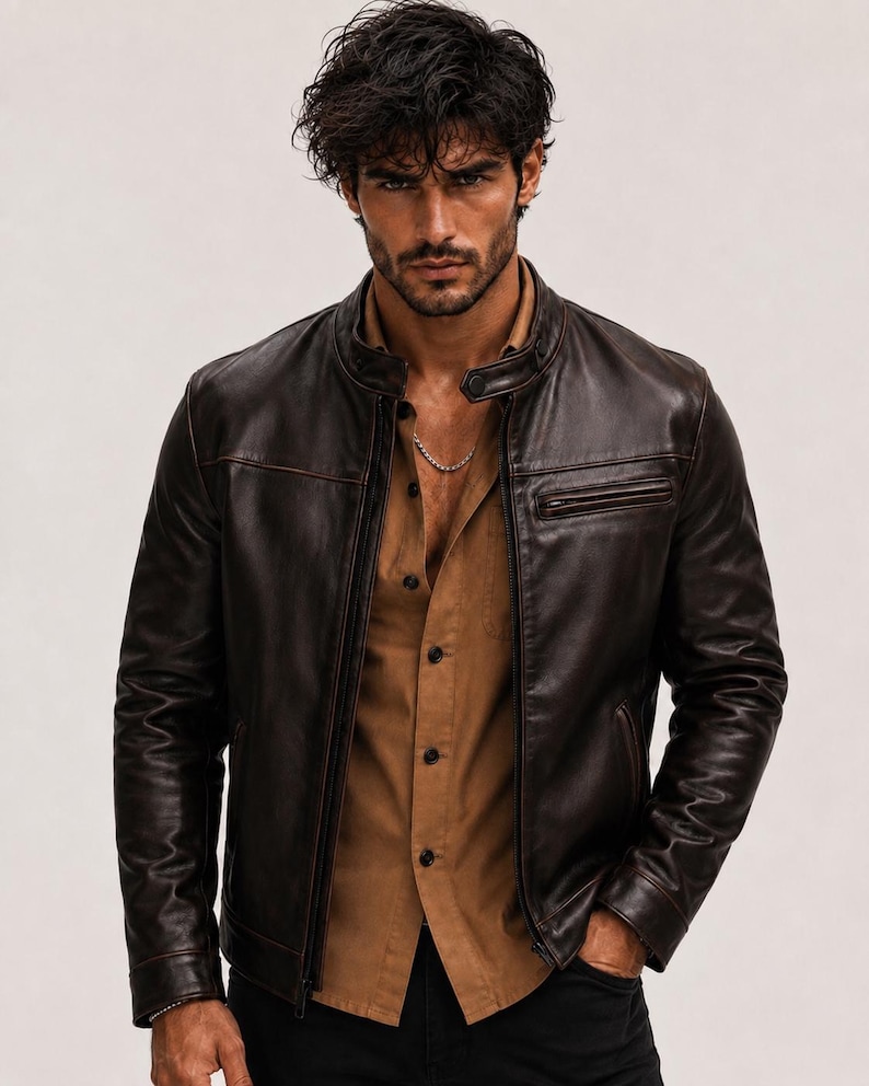 May include: A dark brown leather jacket with a stand-up collar, a front zip fastening, and a zipped chest pocket. The jacket is worn over a tan button-down shirt and black trousers, featuring a silver chain necklace around the neck area.