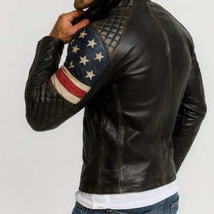Pu&ograve; includere: Giacca in pelle nera con finitura invecchiata. La manica sinistra presenta un design trapuntato e una bandiera americana. La giacca ha un collo alto e una chiusura con cerniera. Il modello indossa una maglietta bianca e jeans blu.