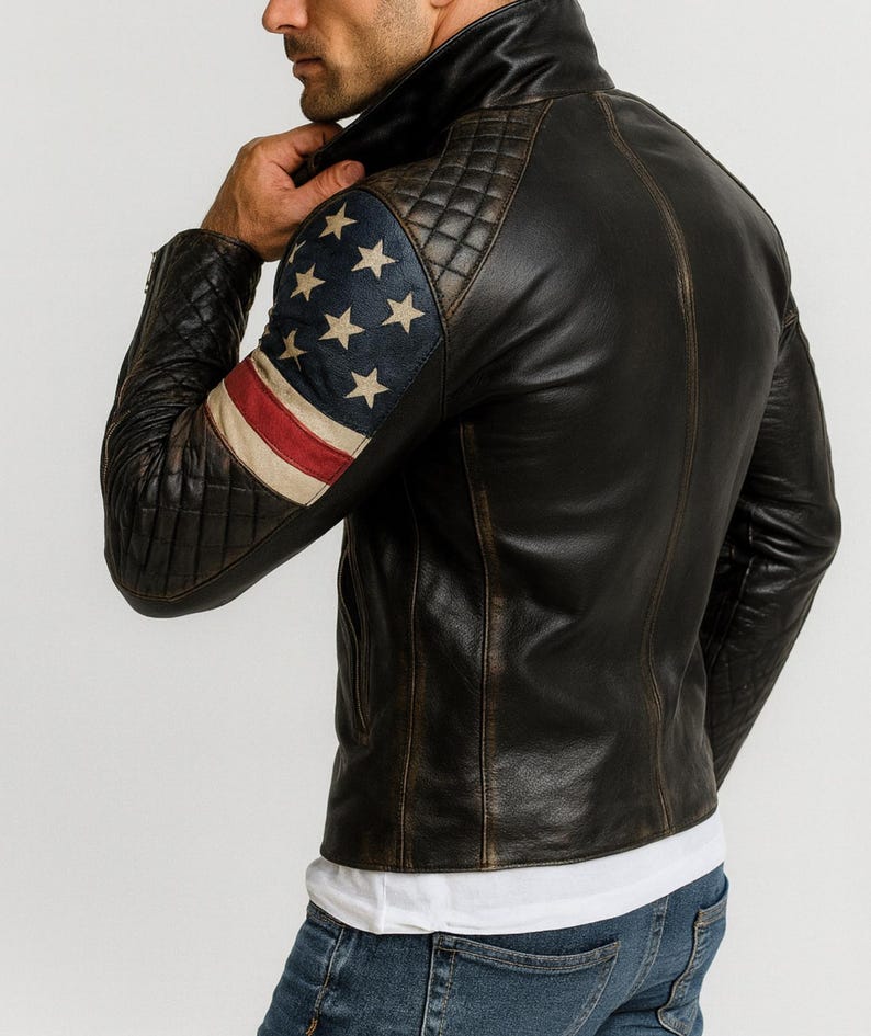 Pu&ograve; includere: Giacca in pelle nera con finitura invecchiata. La manica sinistra presenta un design trapuntato e una bandiera americana. La giacca ha un collo alto e una chiusura con cerniera. Il modello indossa una maglietta bianca e jeans blu.