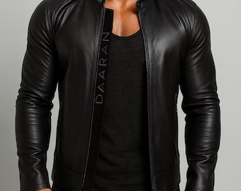 Puede incluir: Chaqueta de cuero negra con cuello alzado y cremallera. La chaqueta tiene un corte entallado y mangas largas. La palabra "DAARAN" está impresa verticalmente en el interior de la chaqueta. El modelo lleva una camiseta negra y vaqueros negros.
