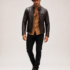 May include: A dark brown leather jacket with a stand-up collar and zippered chest pocket, worn over a tan button-down shirt and black trousers. The outfit is completed with dark brown leather boots, creating a layered look with neutral tones.