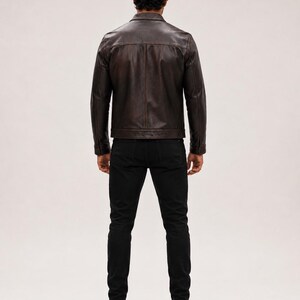 May include: A dark brown leather jacket with a classic collar, long sleeves, and a straight hemline, paired with slim-fit black trousers and dark brown leather boots, viewed from the back against a plain, neutral-coloured background.