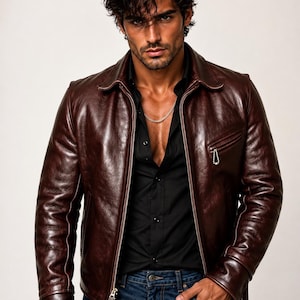 May include: A dark brown leather jacket with a smooth, polished finish, featuring a front zipper closure, a pointed collar, and a small zippered chest pocket. The jacket is worn over a black button-down shirt and dark blue denim jeans.