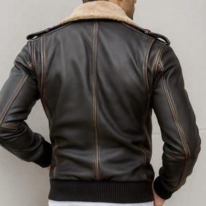 Puede incluir: Chaqueta de cuero marr&oacute;n oscuro con cuello de borrego beige. La chaqueta tiene dobladillo y pu&ntilde;os de canal&eacute;, con detalles desgastados a lo largo de las costuras y bordes. Vista trasera con charreteras.