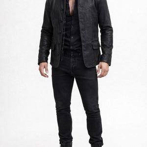 May include: A black leather jacket with a button-up front, paired with a black button-down shirt and black jeans. The outfit is completed with black lace-up boots. The clothing is all black, creating a monochromatic look.