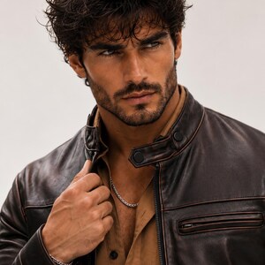 May include: A dark brown leather jacket with a stand-up collar, black snap button closures, and a zippered chest pocket. The jacket is worn over a tan button-down shirt, and a thin silver chain necklace and a matching silver chain bracelet are visible.