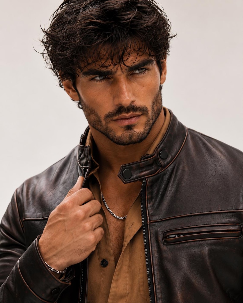 May include: A dark brown leather jacket with a stand-up collar, black snap button closures, and a zippered chest pocket. The jacket is worn over a tan button-down shirt, and a thin silver chain necklace and a matching silver chain bracelet are visible.