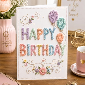 May include: A white greeting card with the text HAPPY BIRTHDAY spelled out in colorful buttons. The card features embroidered floral swirls and three balloon shapes made of buttons in purple, teal, and orange. The border is stitched in a dashed line.