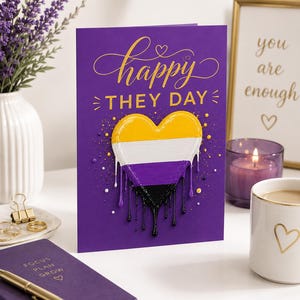 May include: A purple greeting card with gold script that reads "happy THEY DAY." A heart-shaped design in yellow, white, purple, and black is featured. The card is on a white surface with a candle, a mug, and a notebook.