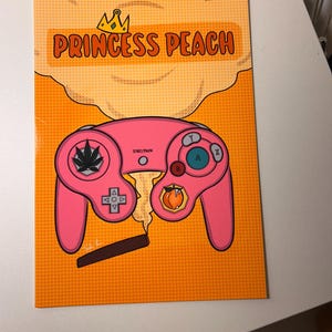 May include: A pop art style print featuring a pink video game controller with a marijuana leaf and a peach. The text "PRINCESS PEACH" is displayed above the controller in orange letters with a gold crown. The background is orange with a dotted pattern.