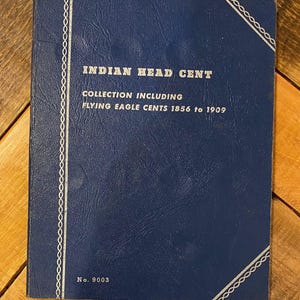 Puede incluir: Un álbum azul de colección de monedas titulado "Indian Head Cent" con el texto "Collection Including Flying Eagle Cents 1856 to 1909". El álbum tiene un borde blanco decorativo y el número "No. 9003" en la parte inferior.