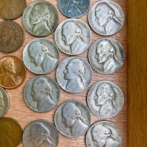 May include: A collection of vintage U.S. coins, including nickels and pennies, displayed in a wooden box. The nickels feature a profile of Thomas Jefferson and the inscription "In God We Trust" and the word "Liberty".