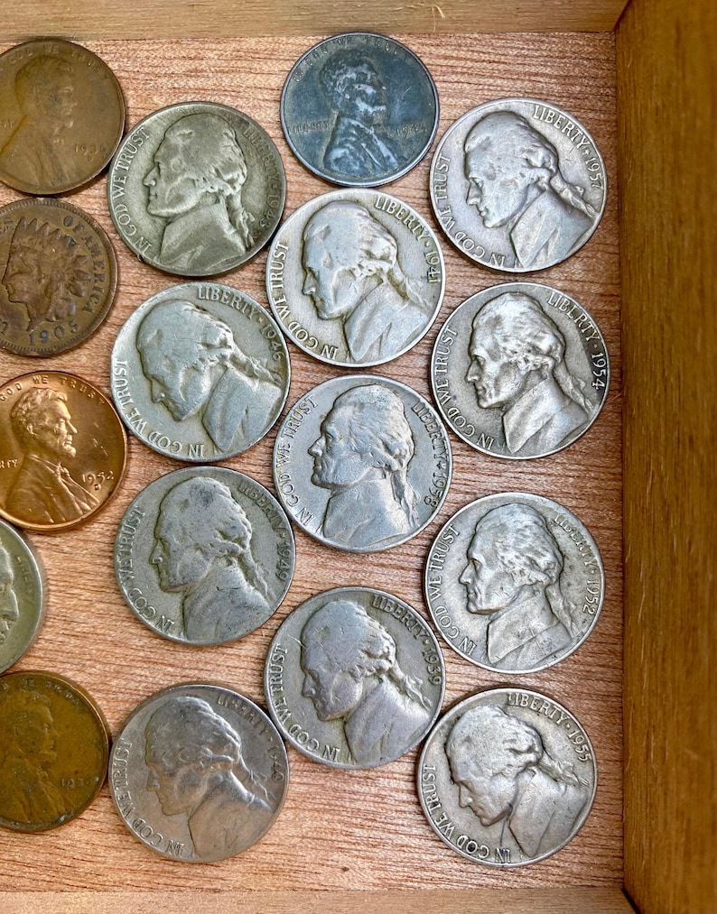 May include: A collection of vintage U.S. coins, including nickels and pennies, displayed in a wooden box. The nickels feature a profile of Thomas Jefferson and the inscription "In God We Trust" and the word "Liberty".