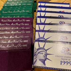 May include: A collection of United States Mint Proof Sets from various years, including 1992, 1993, 1994, and 1999. The sets are in their original packaging, with green, purple, and white boxes. The 1999 set features the Statue of Liberty.