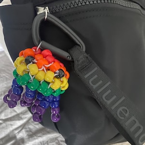 May include: A colorful beaded charm in a rainbow of colors, including red, orange, yellow, green, blue, and purple. The charm is attached to a black carabiner clip, which is connected to a black bag. The charm is a fun accessory.