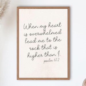 Peut inclure: Une affiche murale rectangulaire dans un cadre en bois brun clair avec un texte noir en écriture cursive sur fond crème qui dit : When my heart is overwhelmed lead me to the rock that is higher than I. psalm 61:2. L'affiche est centrée.