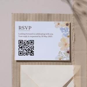 May include: RSVP card with floral accents. The card reads "Looking forward to celebrating with you. Your reply is requested by 20 May 2027." A QR code is present. The card is on a textured background.