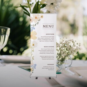 May include: A roll-up menu with floral accents. The menu features a list of dishes, including an appetizer, main course, and dessert. The menu is decorated with watercolor-style flowers in shades of yellow, white, and blue. The menu is displayed on a table setting.
