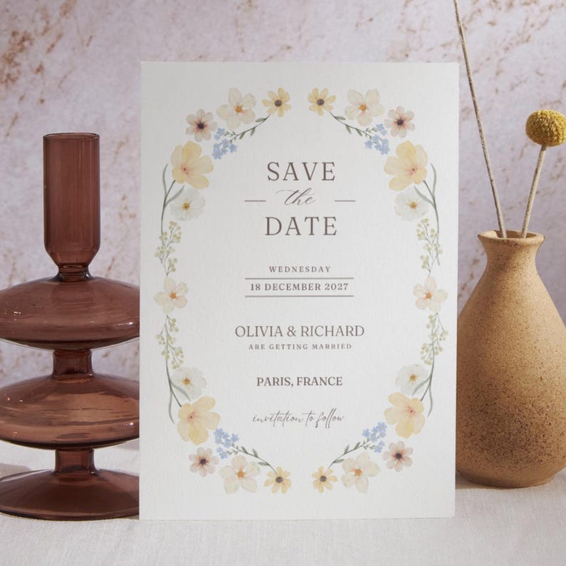 May include: A cream-colored wedding invitation with floral accents. The text reads "Save the Date" with the names Olivia & Richard, the date December 18, 2027, and location Paris, France. The invitation is surrounded by a wreath of flowers.