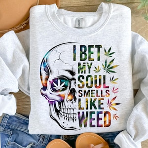 Peut inclure: Sweat-shirt gris clair avec un motif de crâne. Le crâne est multicolore avec une feuille de cannabis dans l'orbite de l'œil. Le texte sur le sweat-shirt se lit "I BET MY SOUL SMELLS LIKE WEED" avec des feuilles de cannabis.