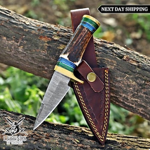 May include: A Damascus steel knife with a patterned blade and an antler and brass handle. The handle has blue and green bands. The knife is in a brown leather sheath with decorative stitching.