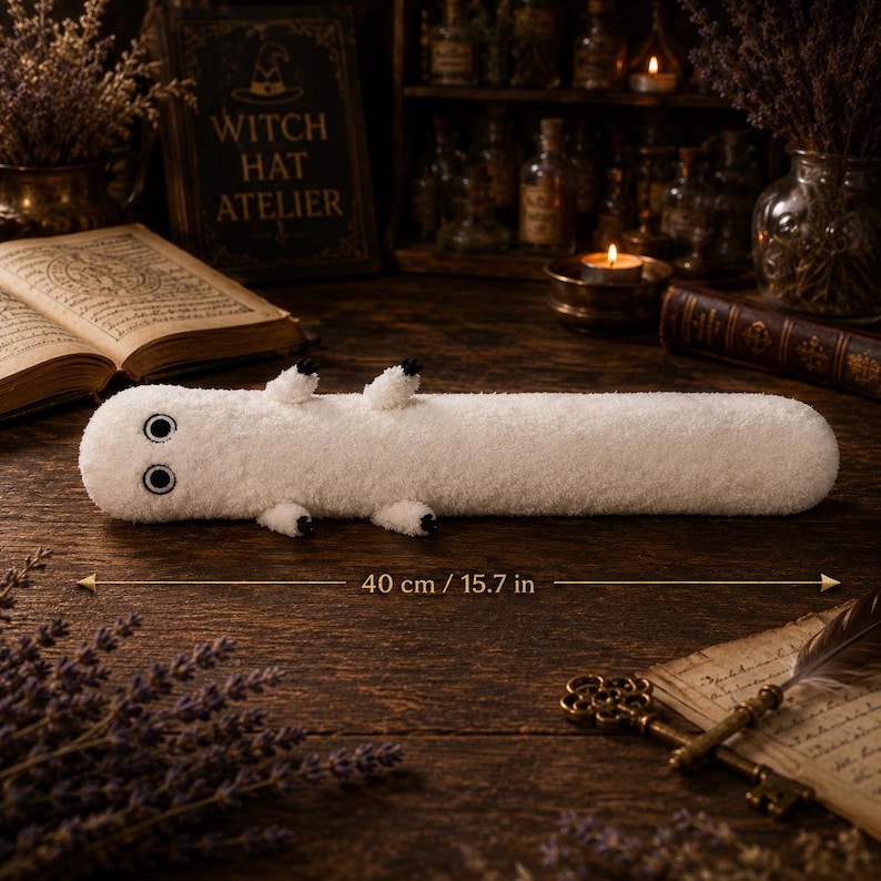 May include: A long, cylindrical white plush toy with a fuzzy texture, two large black and white eyes, and four small limbs with black tips. The toy is positioned horizontally and features a measurement indicator showing a length of 15.7 inches.